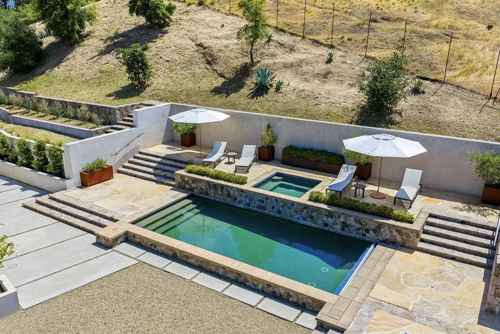 Patio with a swimming pool, hot tub, sun loungers with umbrellas, potted plants, and stairs leading to a grassy hillside with trees and desert plants.