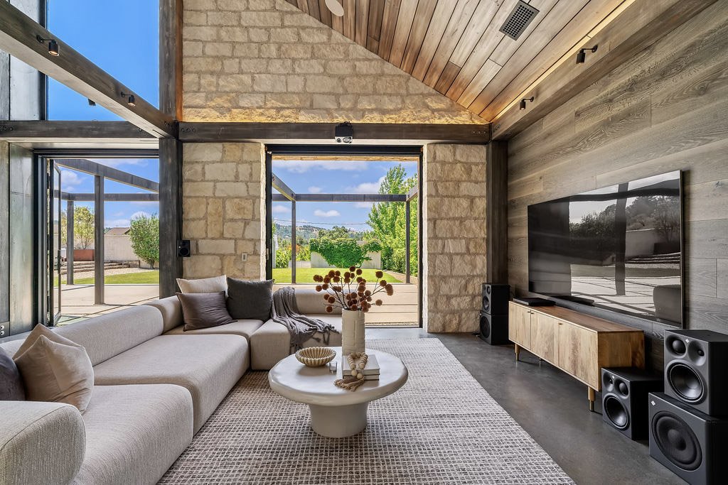 Modern living room with large windows, stone and wood walls, and a beige sectional sofa.