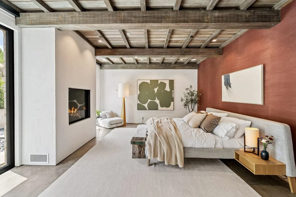 Modern living room with a bed, placed against a painted red accent wall, and a white shelf with a lamp and flowers. There is a fireplace on the white wall, a large window on the left, and artwork on the walls, with a visible ceiling featuring exposed