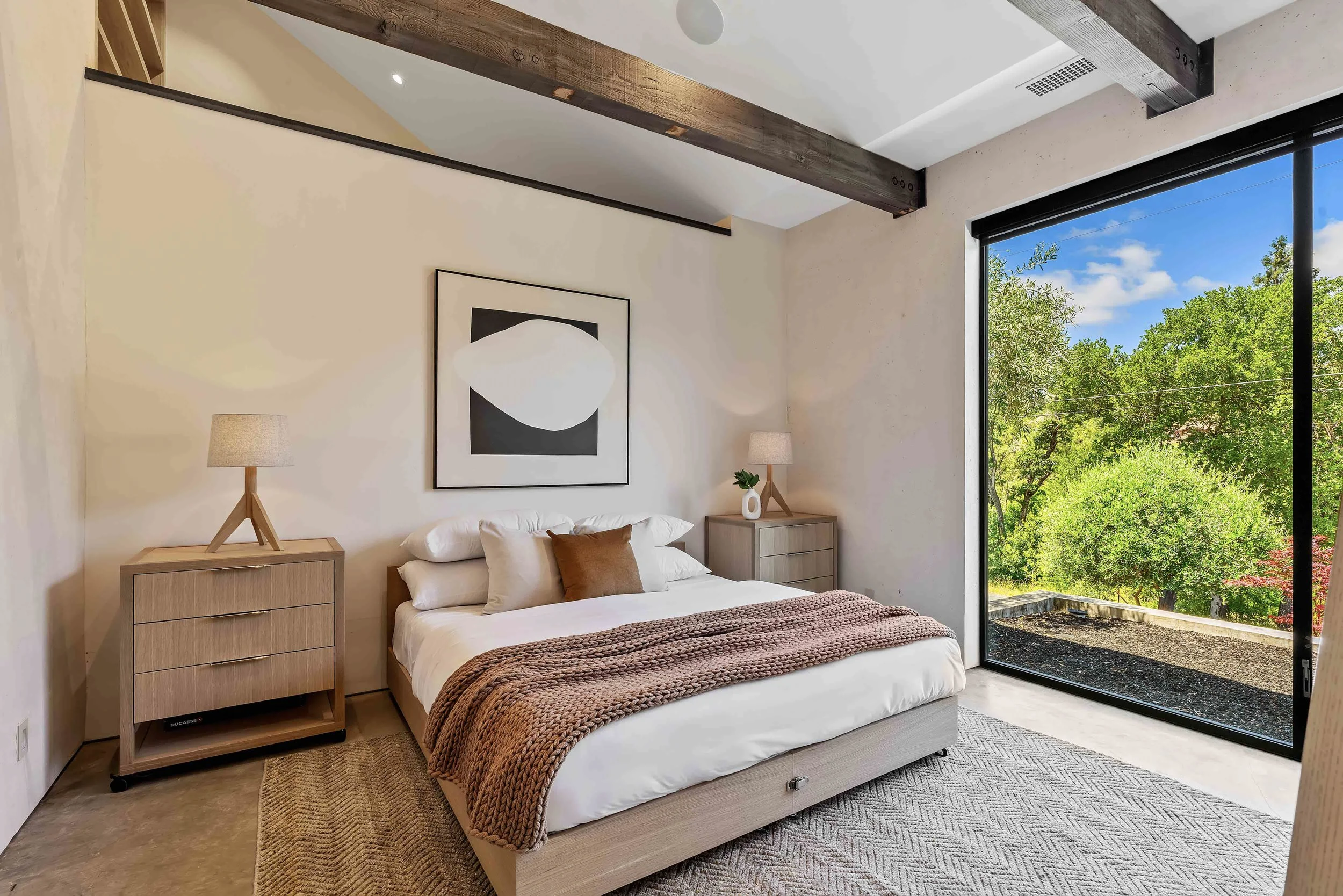 Bedroom with a large window showing green trees and blue sky outside. The bed has white bedding and a brown knitted throw, with brown and white pillows. Two wooden nightstands with matching lamps are on either side of the bed. A black and white abstr