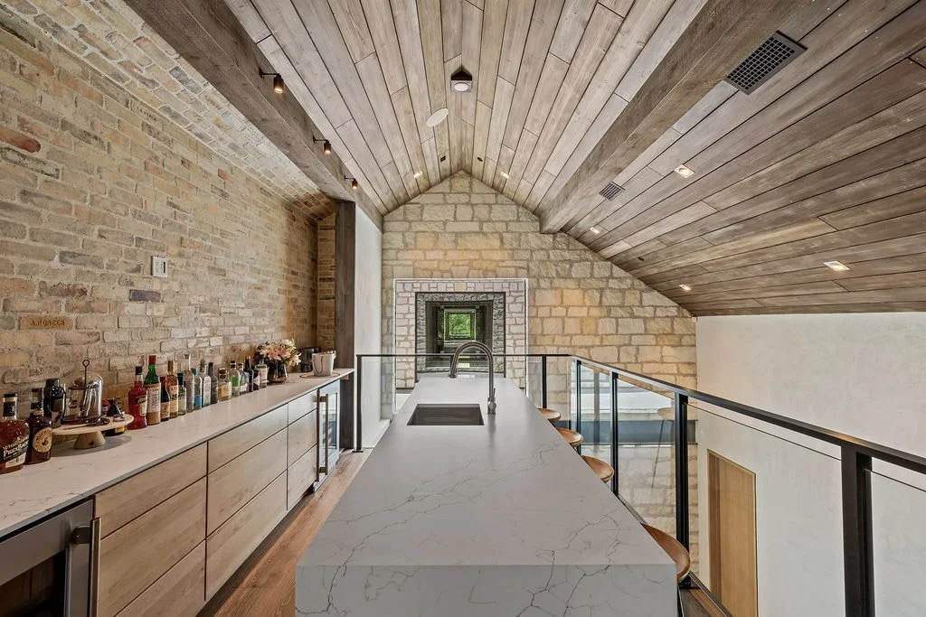 A modern kitchen with a marble island, wooden ceiling, brick walls, and bar stools.