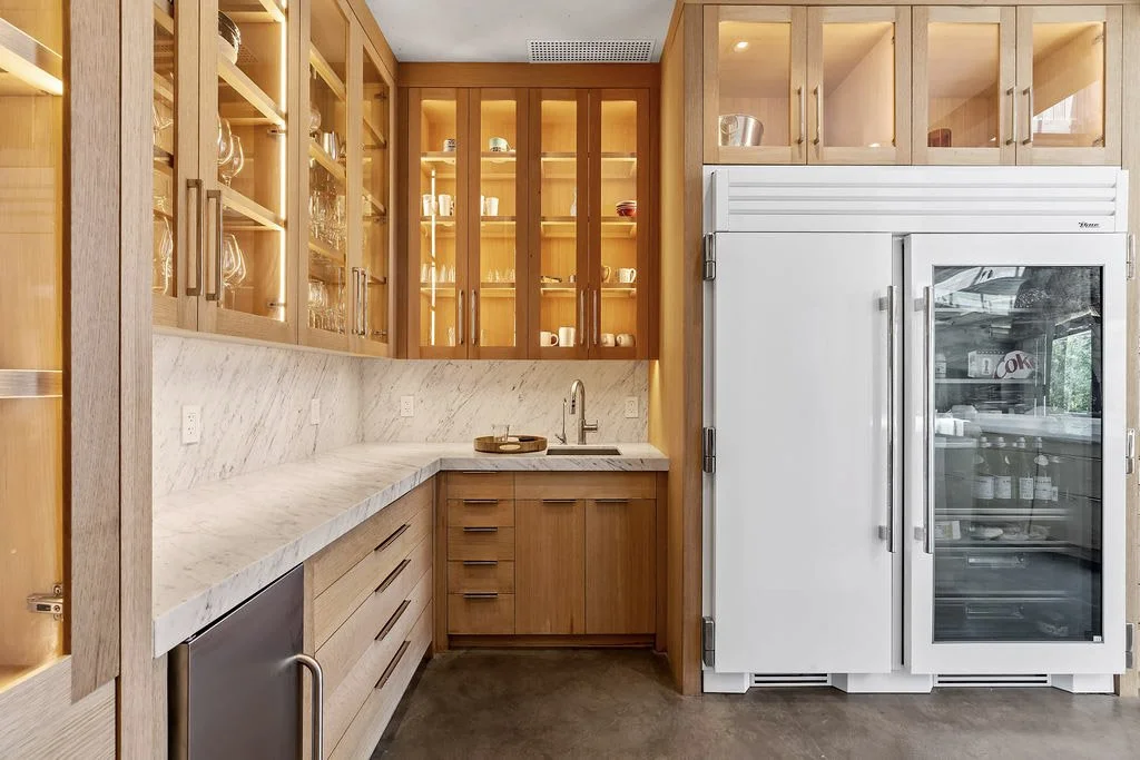 Kitchen with wooden cabinets, white marble countertops, and a white refrigerator with a glass door freezer. The upper cabinets have glass doors displaying dishes and glassware.