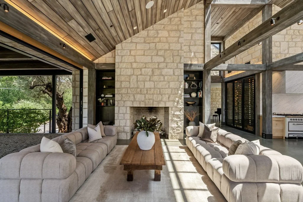 Living room with beige sofas, a wooden coffee table with a potted plant, a stone fireplace, built-in shelves, large windows, and a wooden ceiling.
