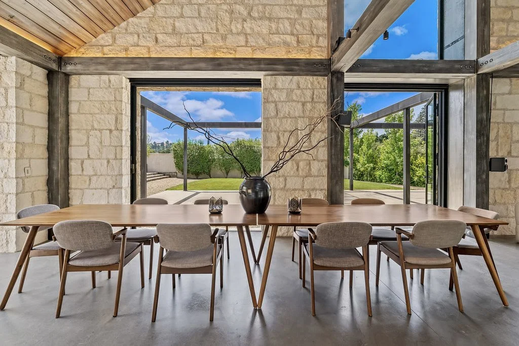 Modern dining room with tan brick walls, large windows showing outdoor greenery, a wooden table, and eight upholstered chairs, with a black vase and decorative candles on the table.