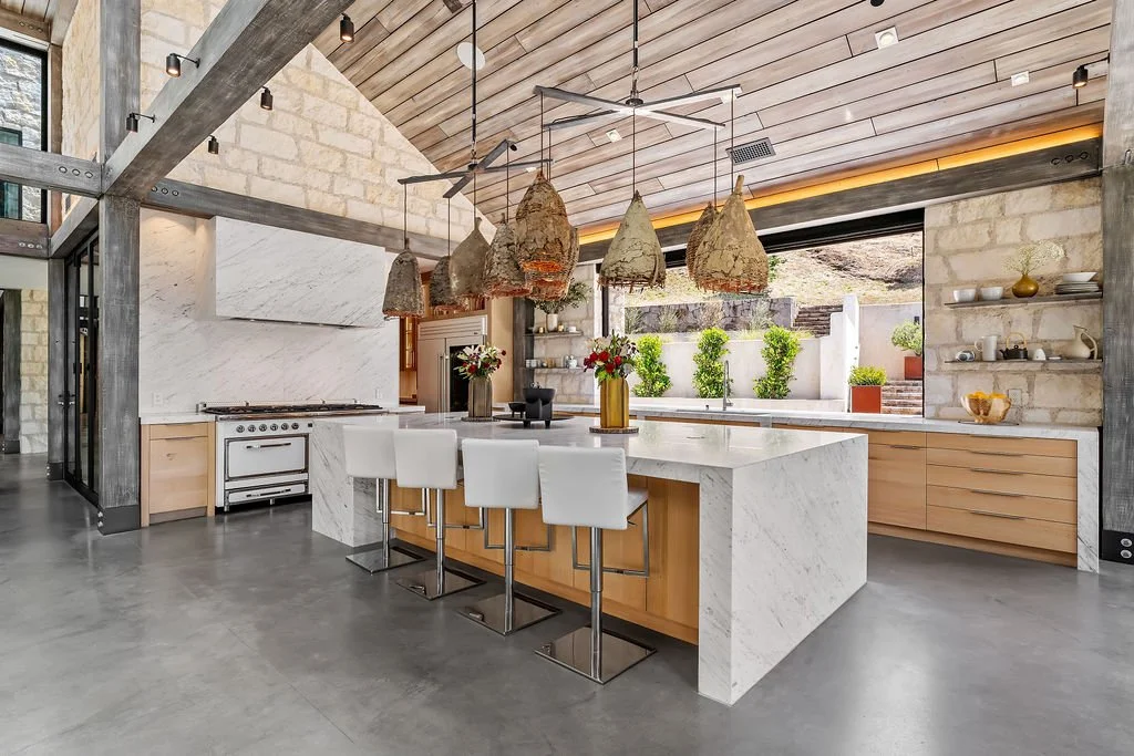 Modern kitchen with large white marble island, four white barstools, built-in stove, open shelves with dishes, large windows with plants outside, wood and stone accents, hanging pendant lights with unique covers, and a wood ceiling.