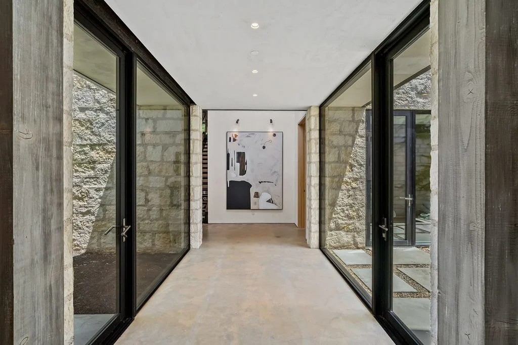 Modern indoor hallway with large glass sliding doors on both sides, stone walls, and an abstract painting on the back wall.