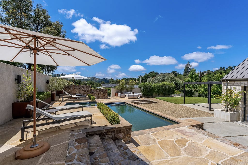 Backyard with a swimming pool, lounge chairs, umbrellas, and green trees under a blue sky with white clouds.