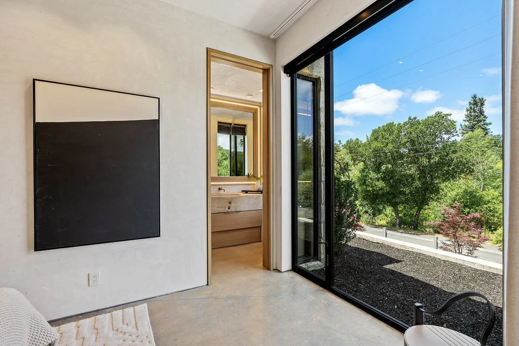 A bright, modern room with a large sliding glass door showing a view of green trees and a blue sky. Inside, there is a minimalist black and white abstract painting on the wall, a wooden-framed mirror, and a bathroom with a stone sink visible through 