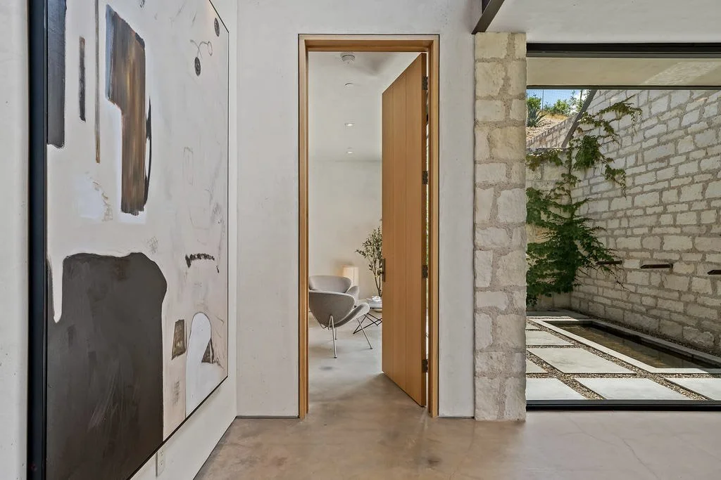 Interior view of a modern space showing an open wooden door leading to a sitting area with white chairs and a table, stone walls with greenery outside, and abstract artwork on the wall.
