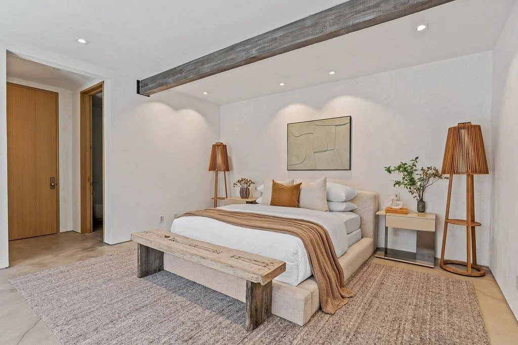 A minimalist bedroom with a beige bed, wooden bench at the foot, two wooden floor lamps, white side table with a potted green plant, beige rug, and abstract wall art. The room has white walls, a ceiling with recessed lighting, and a wooden ceiling be