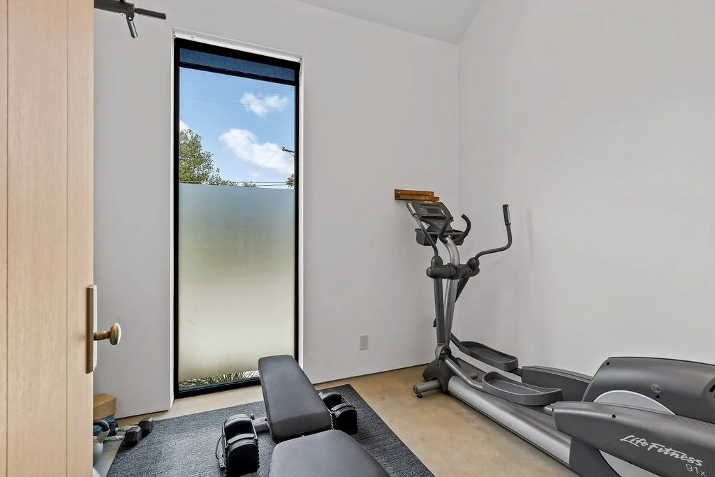 A small home gym with an elliptical machine, adjustable weight bench, dumbbells, and a door with a window showing blue sky and clouds.