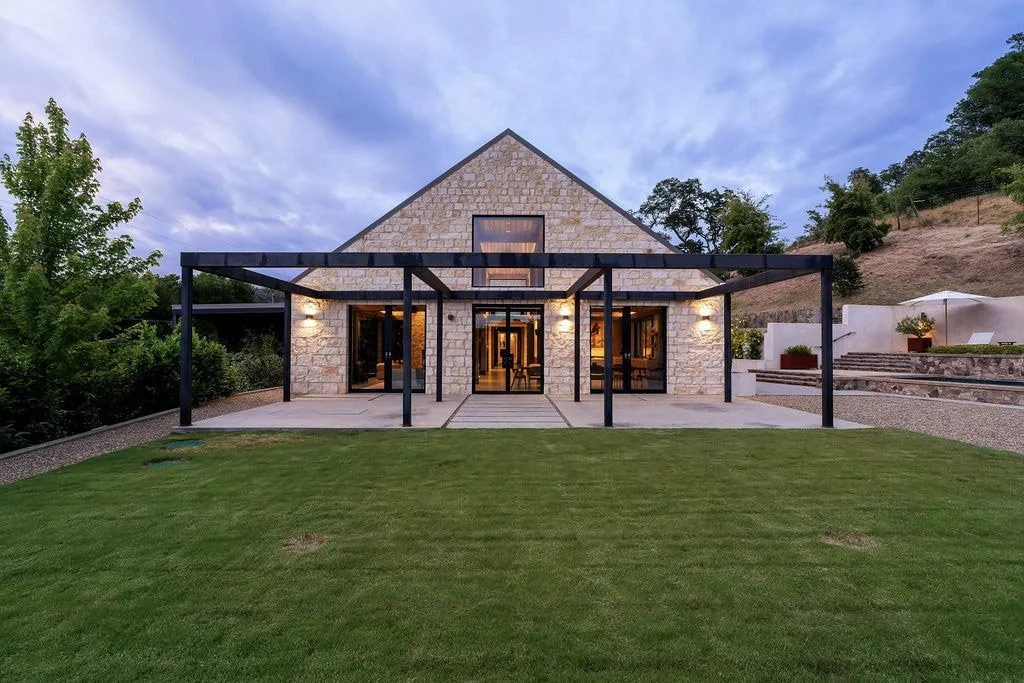 Modern house with stone exterior, black metal pergola, large windows, and a well-maintained lawn, set against a hillside with trees.