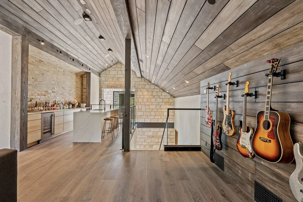 Modern upper-floor recreational space with a slanted wooden ceiling, four hanging guitars on the dark wooden wall, a bar area with bottles and glasses, and a marble-topped kitchen island with barstools.