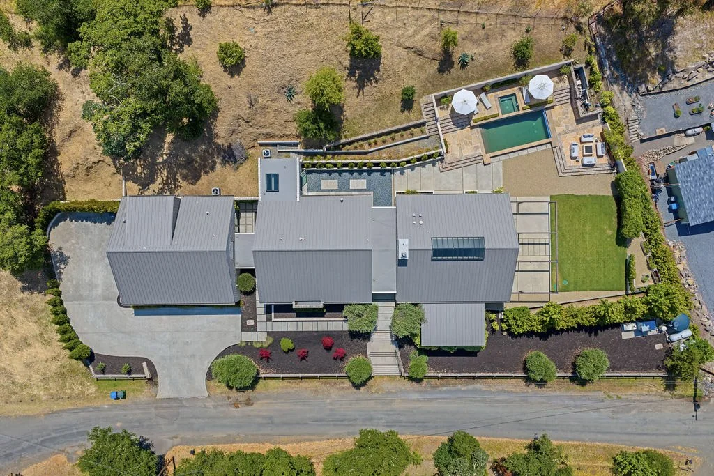 Aerial view of a modern house with a grey metal roof, surrounding landscaped yard with trees, a driveway, and a backyard with a pool, hot tub, and patio area with umbrellas.