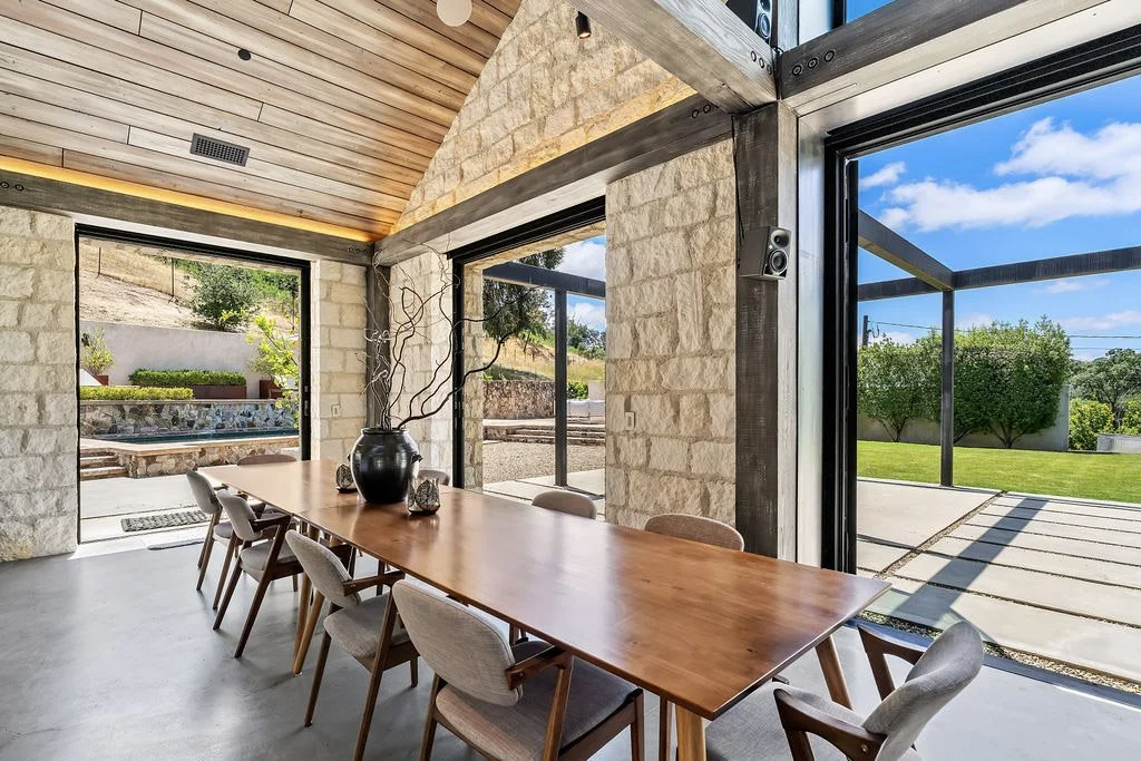 Modern dining room with wood-paneled ceiling, stone walls, large glass sliding doors, and a wooden dining table with chairs, overlooking a landscaped backyard.