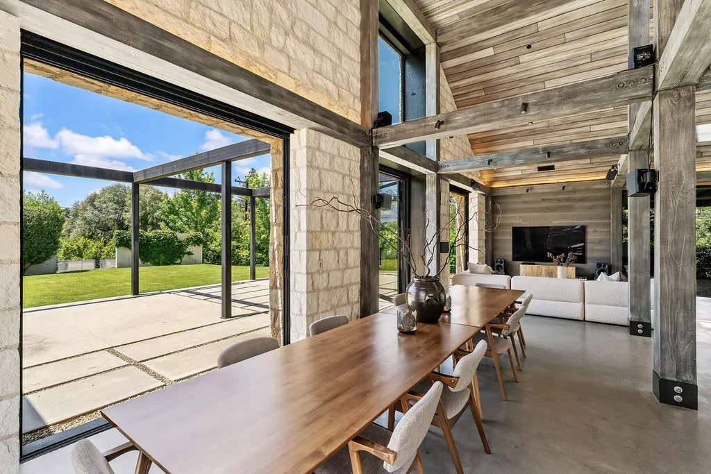 Modern open-concept living and dining area with floor-to-ceiling glass windows, wooden ceiling and beams, stone and wood interior walls, a large wooden dining table with chairs, and a living space with a white sofa and TV, overlooking a lush green ba