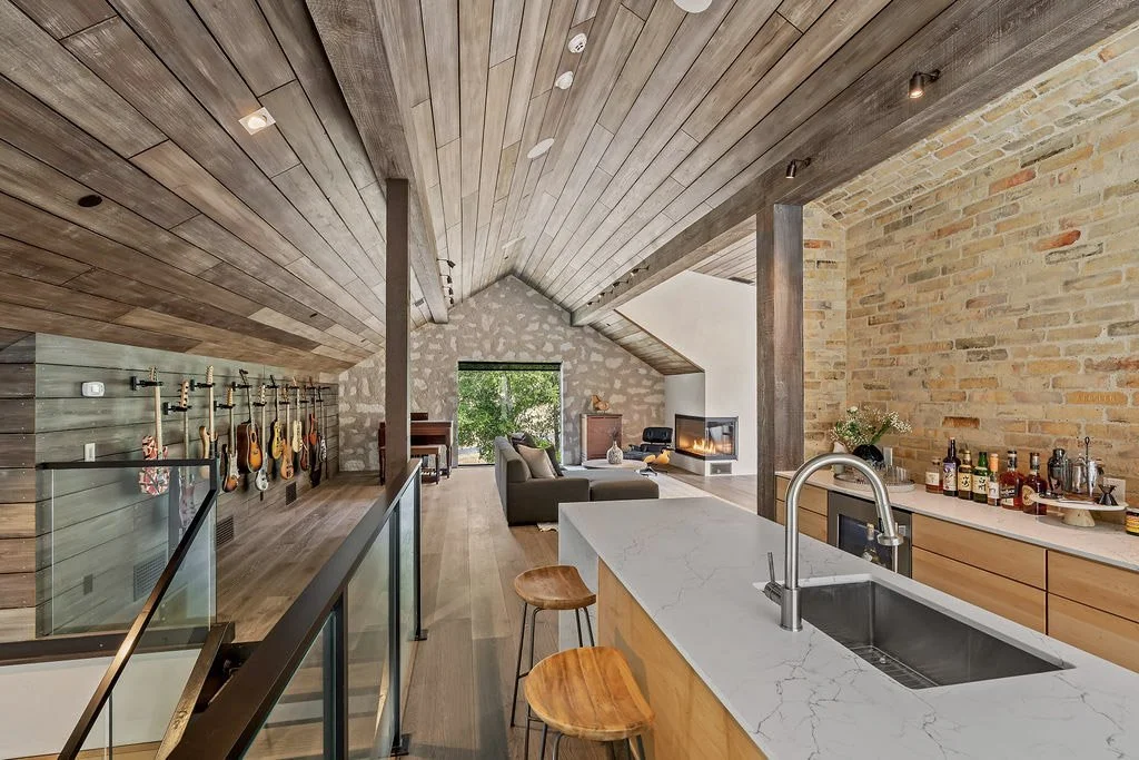 Open-concept living room and kitchen with vaulted wood-paneled ceiling, brick accent wall, and Guitars on a wall display