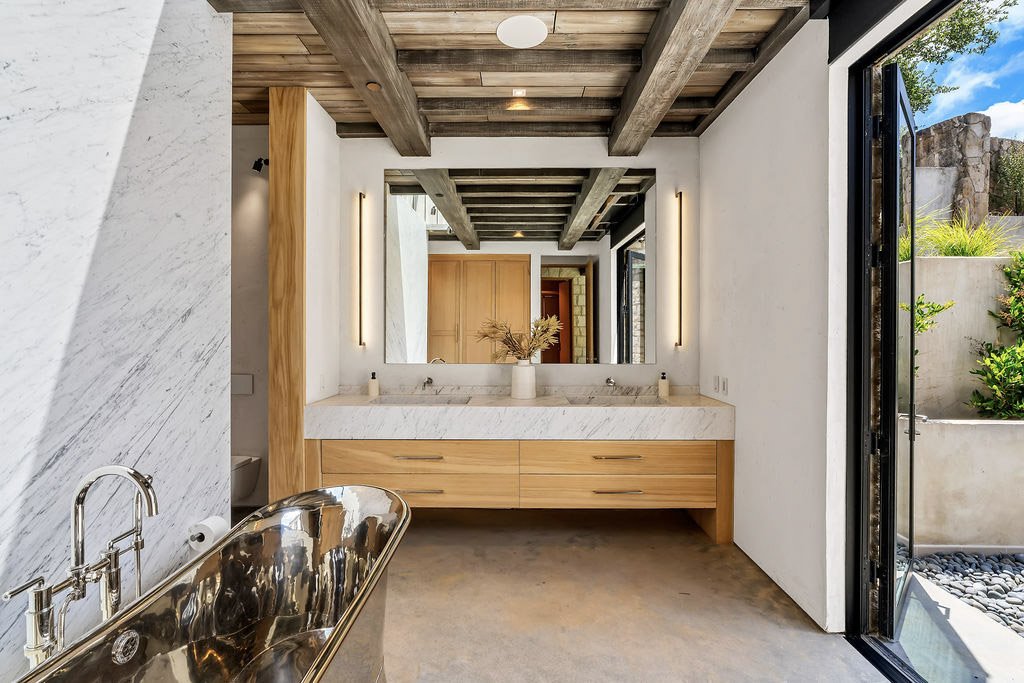 Modern bathroom with wooden accents, a large mirror, and a metal bathtub, with outdoor scenery visible through an open door.