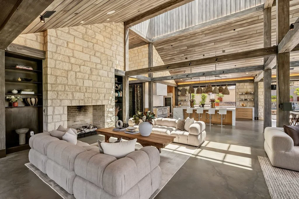 Open-concept living room and kitchen with a rustic modern design, featuring beige sofas, a stone fireplace, wooden accents, and a kitchen island with white barstools.