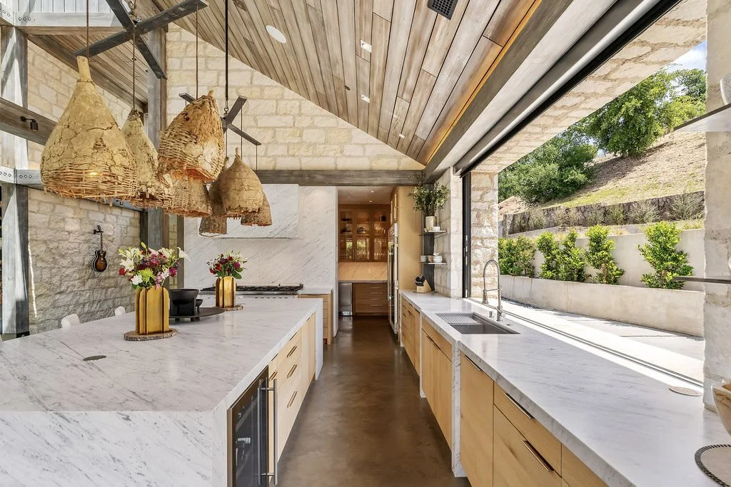 Modern kitchen with large window overlooking a landscaped backyard, featuring white marble countertops, wooden cabinets, and woven pendant lights.