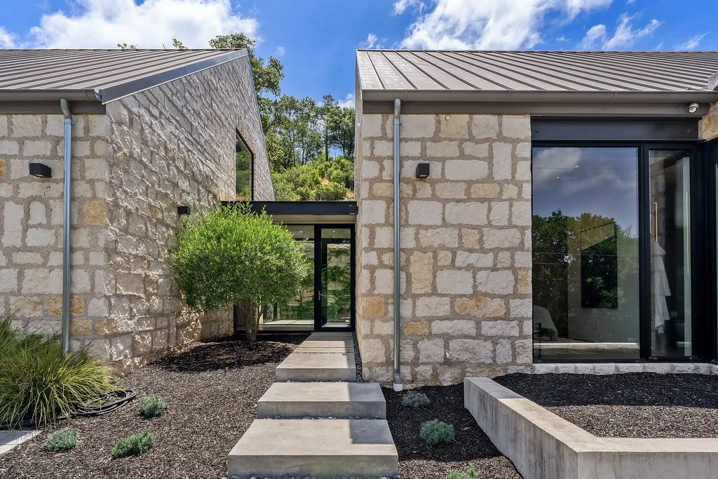 Modern house with stone exterior walls, metal roof, glass sliding door, small landscaped yard with plants and shrubs, and a pathway leading to the entrance.