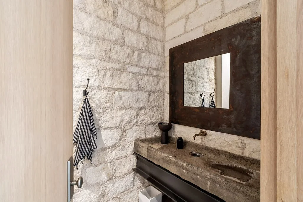 A bathroom with a stone wall, a rectangular mirror with a dark wooden frame, a rustic concrete sink, and a black faucet. A striped towel hangs on a hook near the door.