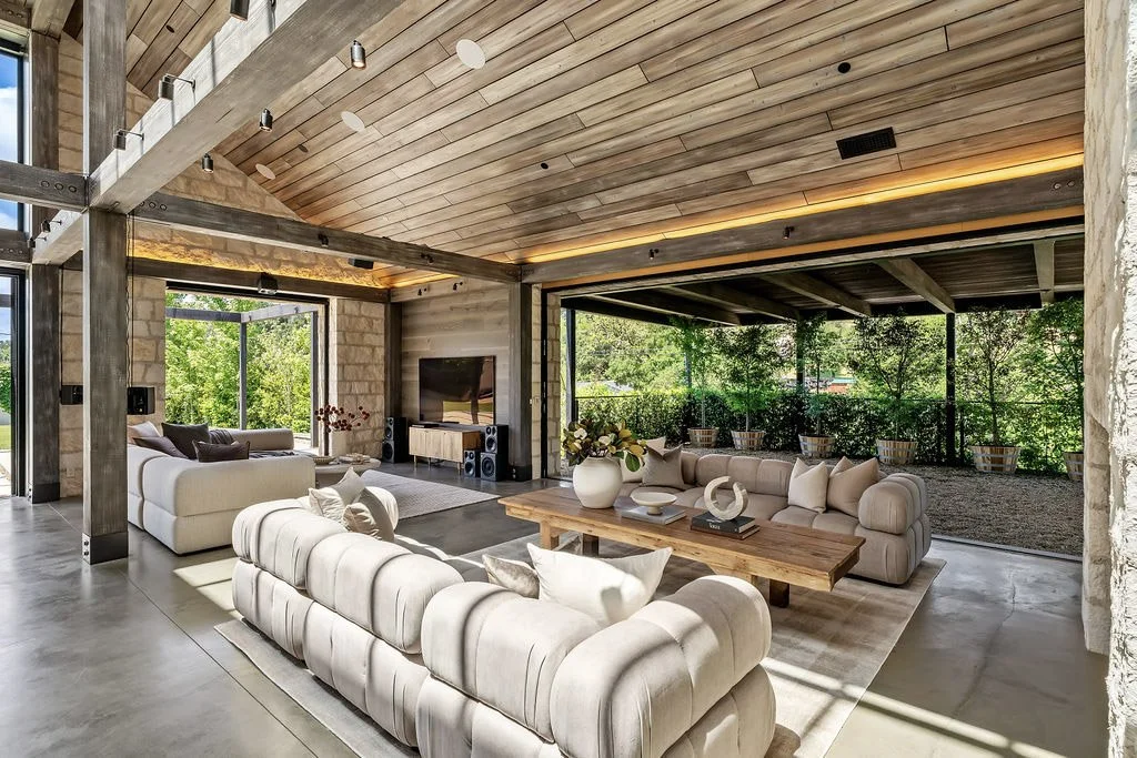 Modern living room with large windows, beige sofas, wooden ceiling, and a view of greenery outside.