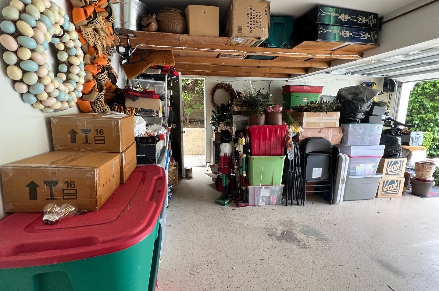 Garage filled with boxes, storage bins, and various household items, including decorations, plants, and tools, with an open garage door showing greenery outside.