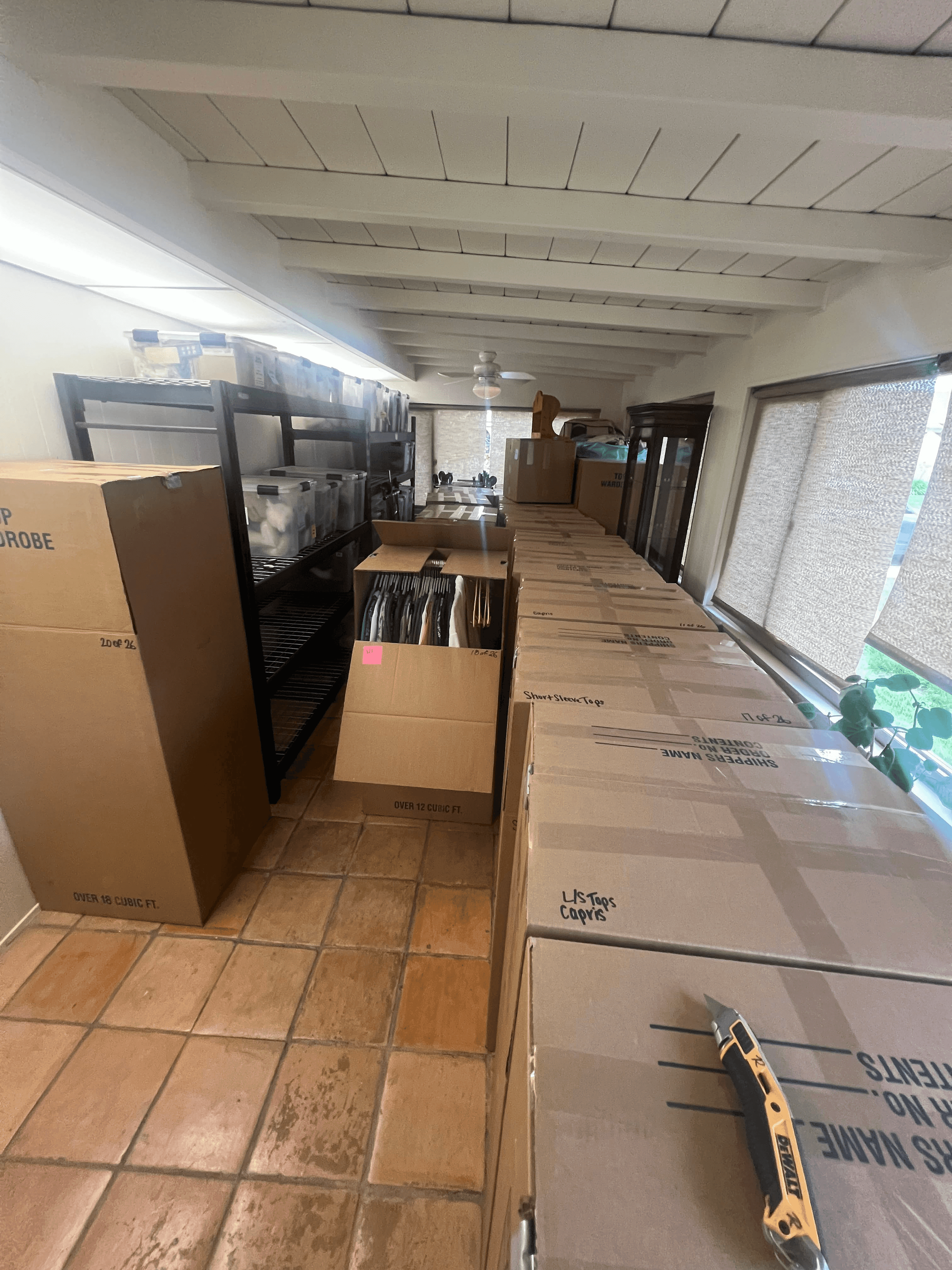 A room filled with cardboard boxes and storage shelves. Some boxes are labeled, and there is a utility knife on a large flat box in the foreground. The space has tiled flooring and blinds covering windows on the right side.