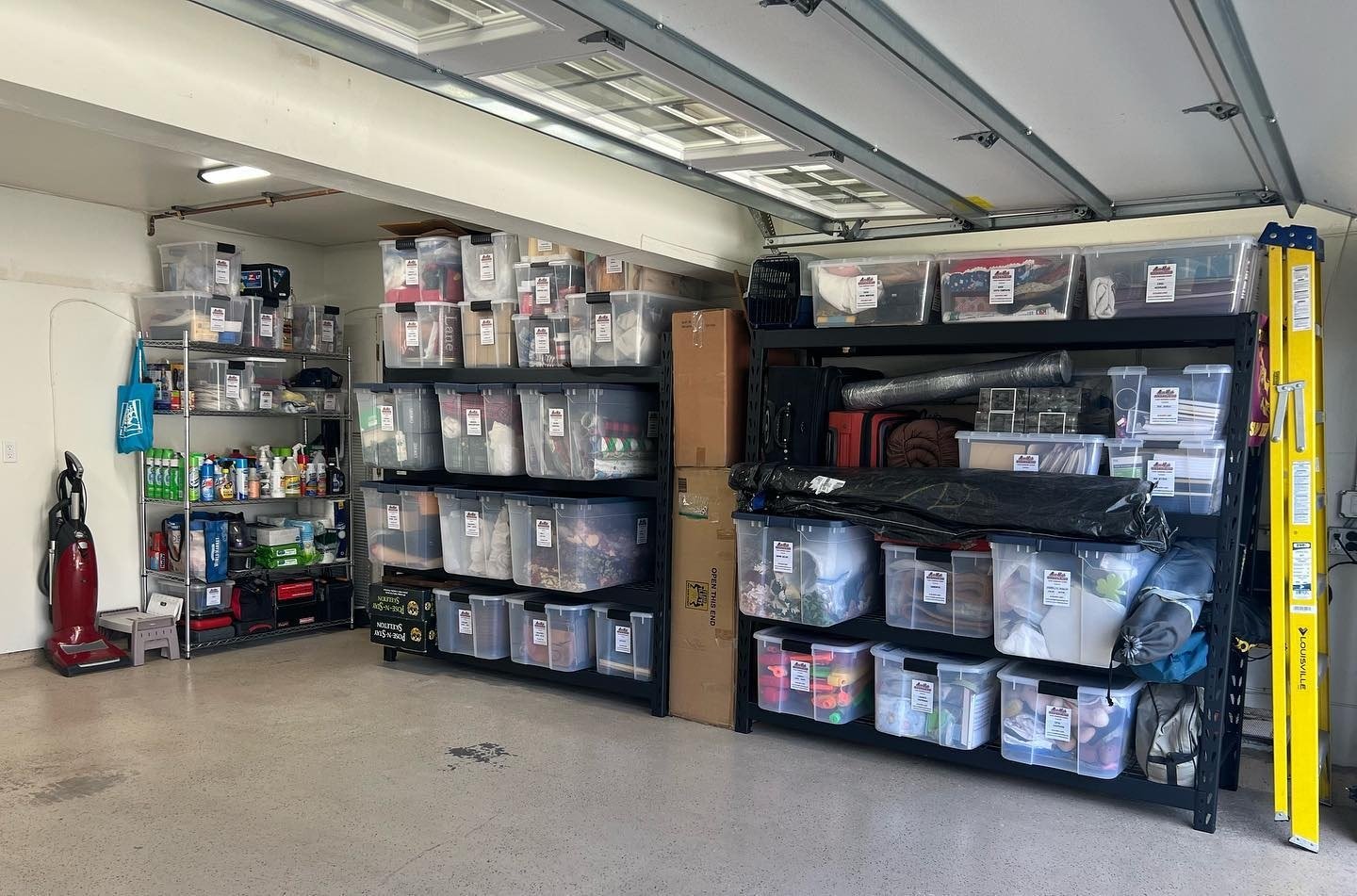 Garage storage with black metal shelves filled with organized clear plastic bins, a metal utility shelf with cleaning supplies, and a yellow ladder on the right side.