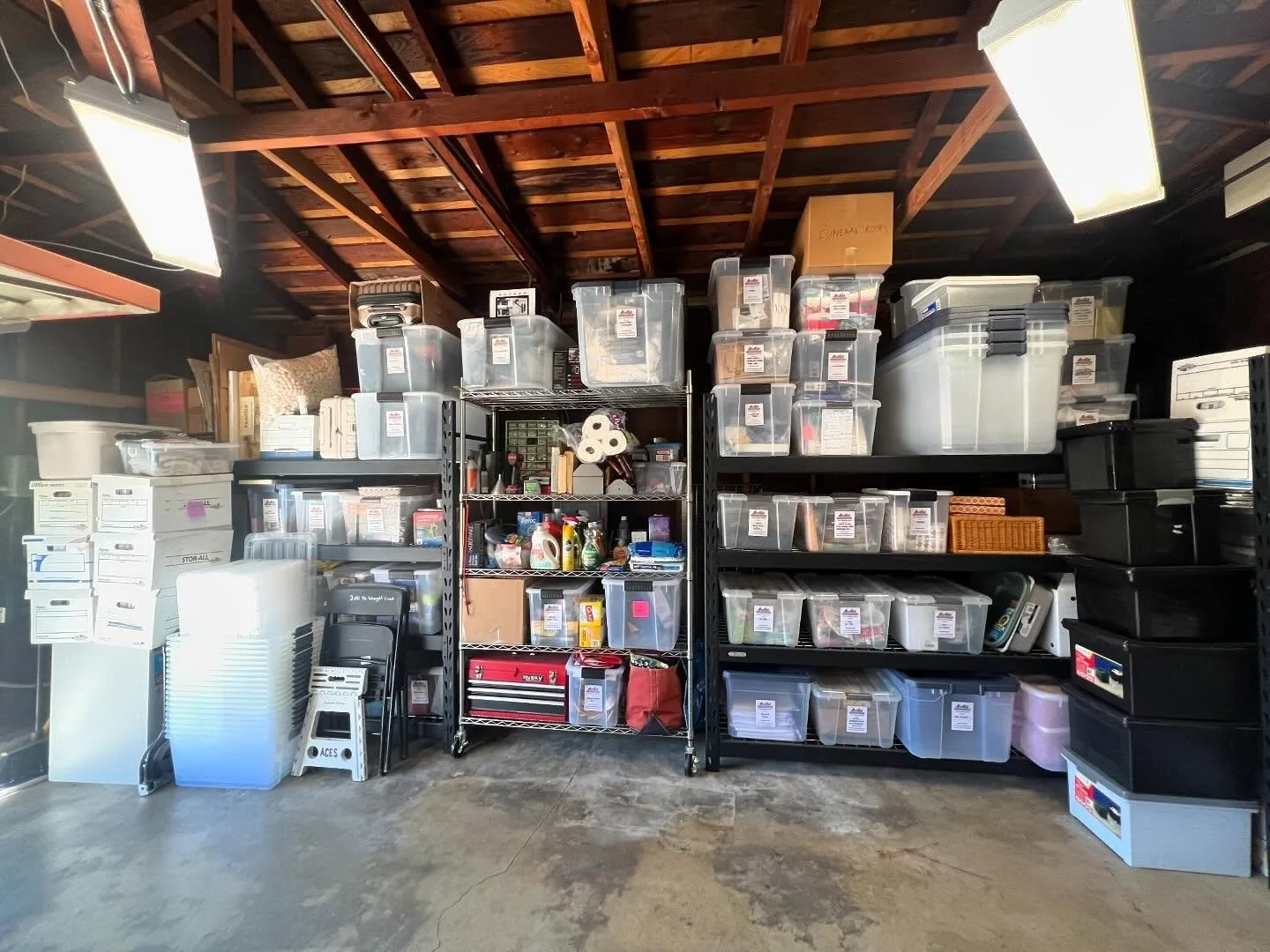A garage storage area with shelves filled with plastic bins containing various items, and cardboard boxes on the left side, with some containers labeled, and a concrete floor.