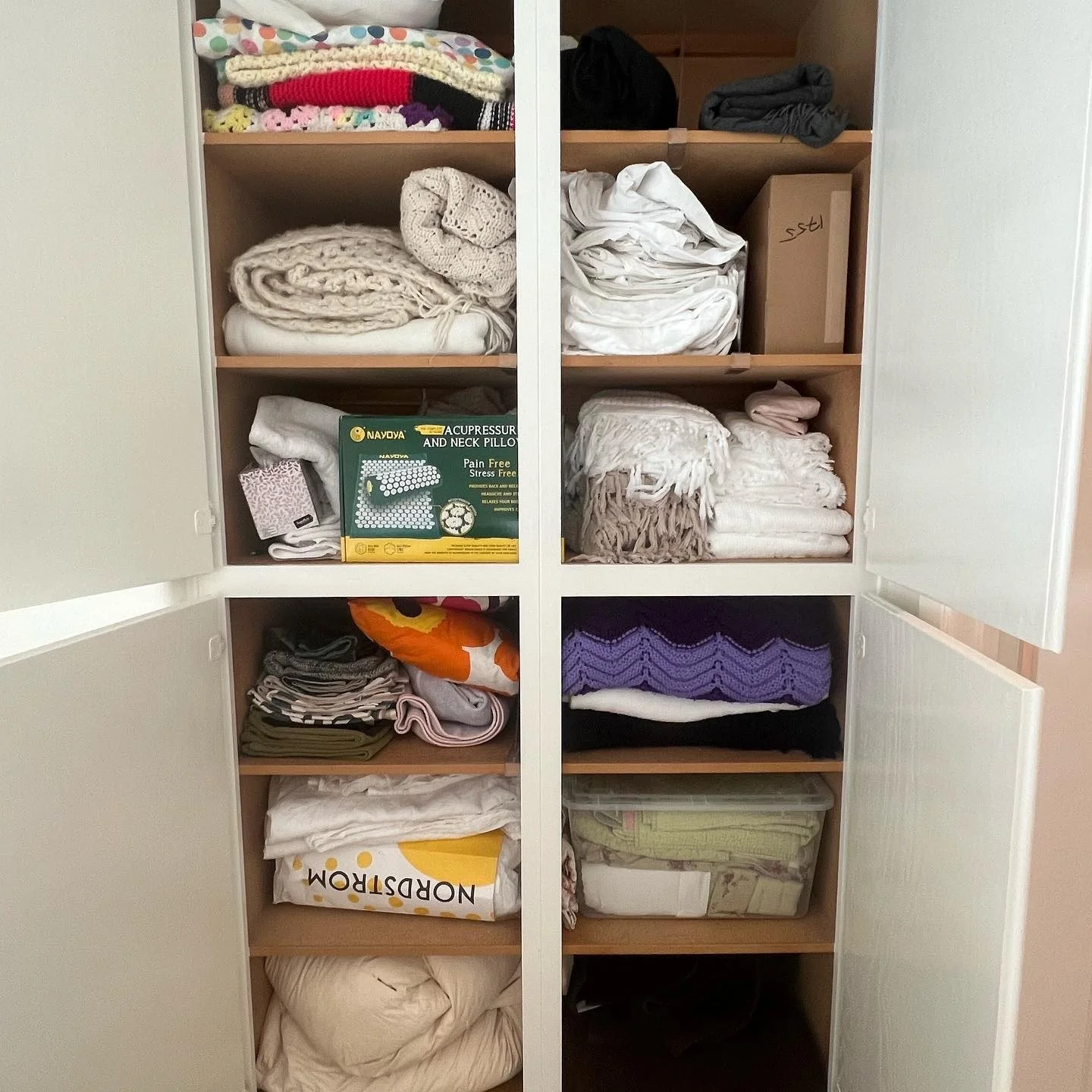 An organized white closet with open doors, containing shelves filled with folded blankets, towels, linens, and other textiles, with a box labeled 'st' and a package of acupressure pillows.