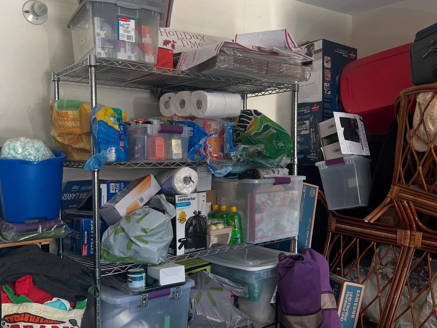 A metal shelving unit filled with various household items, including toilet paper rolls, cleaning supplies, plastic containers, and bags. Beside the shelving, there are stacked chairs and a red suitcase against the wall.