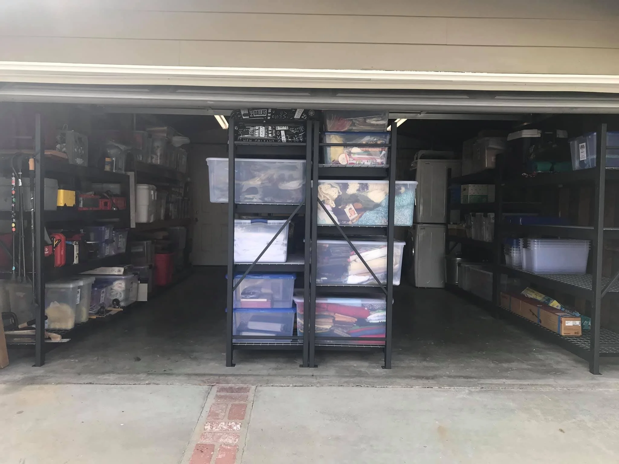 View of a garage with organized shelves and plastic containers filled with various items, visible through the open garage door.