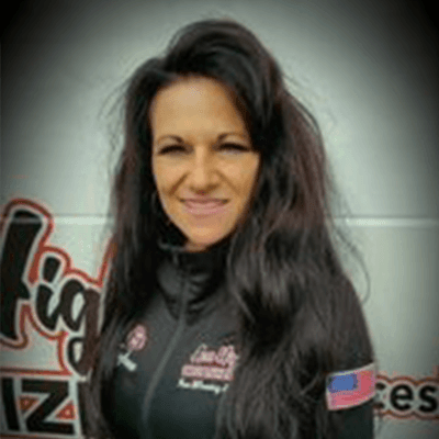 A woman with long dark hair smiling, wearing a black jacket with logos, standing outdoors.