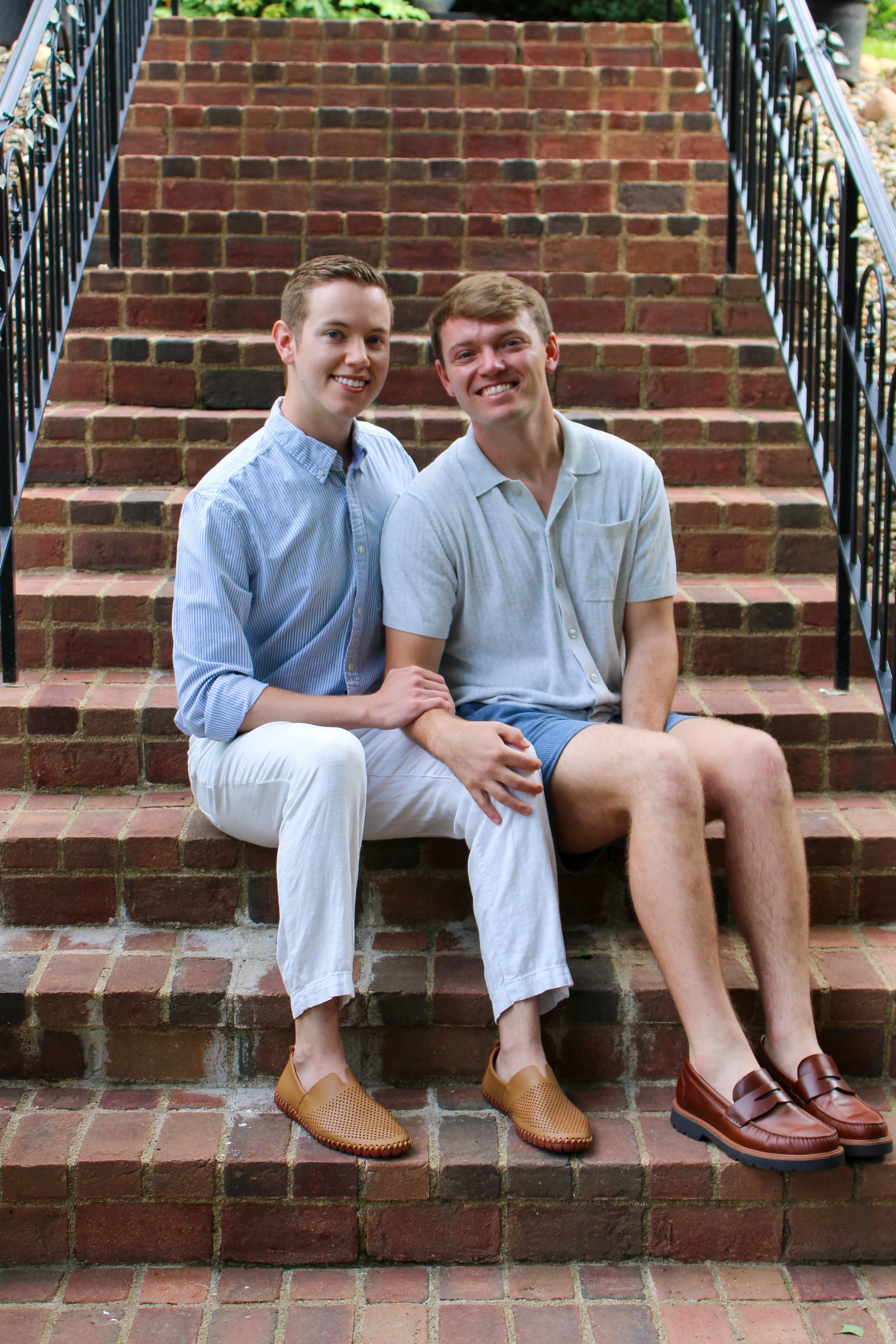 Two young men sitting on brick stairs, smiling, wearing casual summer clothes, with black metal railing on either side.