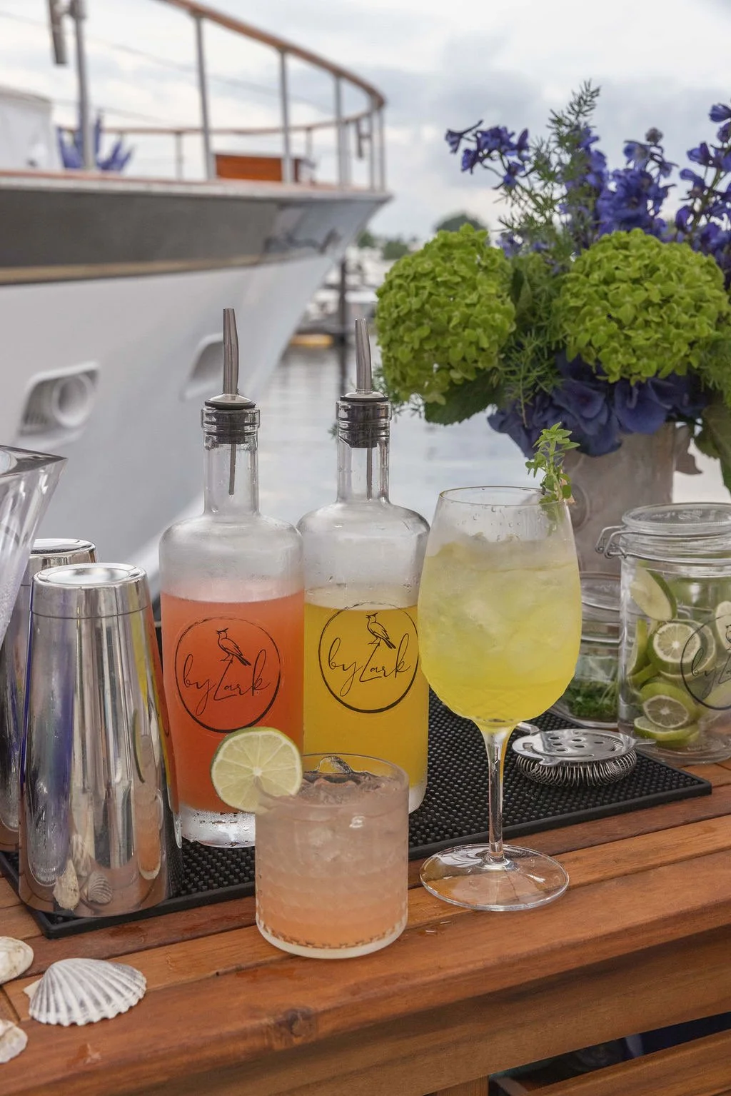 A variety of colorful beverages with lemon slices on a wooden bar table, with a yacht and blue flowers in the background.