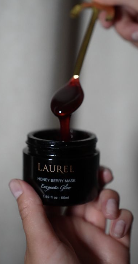 A hand holding a black jar labeled 'Laurel Honey Berry Mask Exquisite Gloss,' with a spoon above the jar containing a dark red gel or honey-like substance.