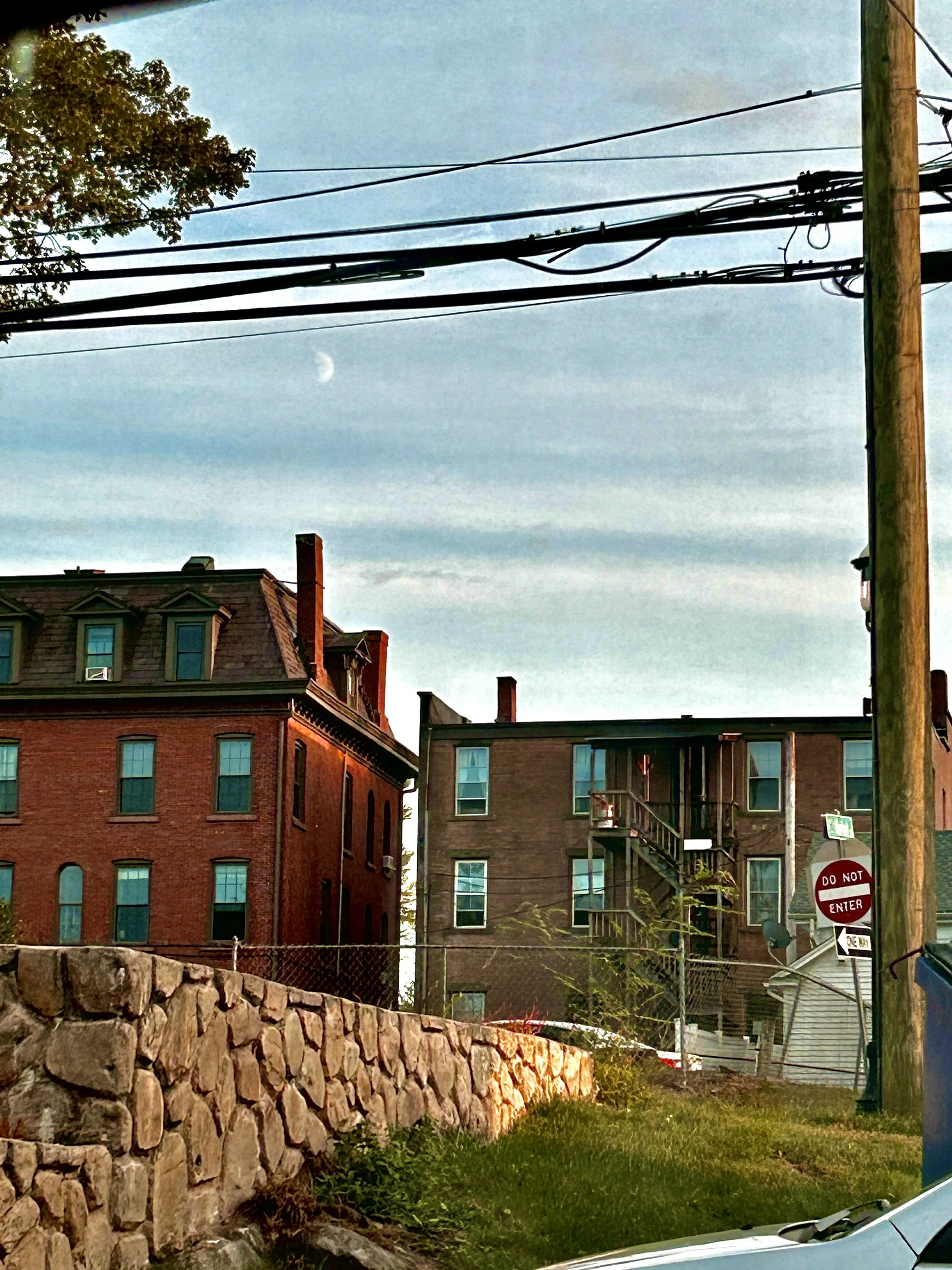 View of a city neighborhood with brick buildings, utility poles, and power lines under a partly cloudy sky with a visible moon.
