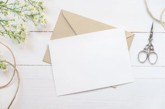 Flat lay of a blank white card on top of a kraft paper envelope, with a pair of scissors, some twine, and a small bunch of white flowers on a white wooden surface.