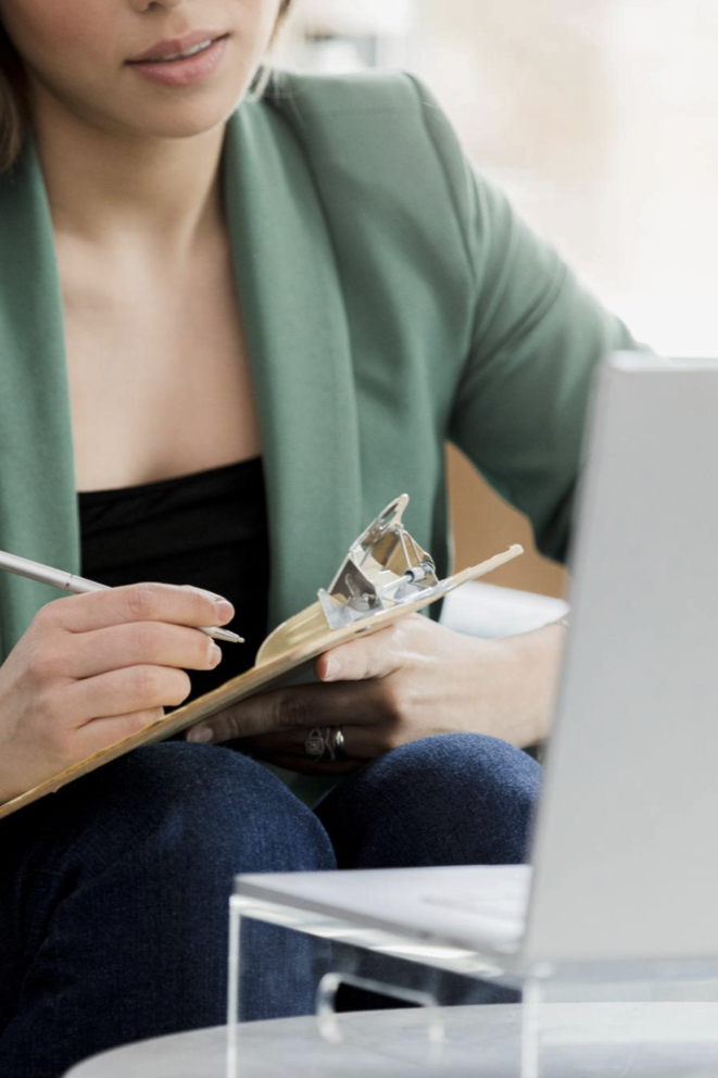 A woman sitting at a desk, near a laptop, holding a clipboard with a mechanical device attached to it, and writing with a pen.