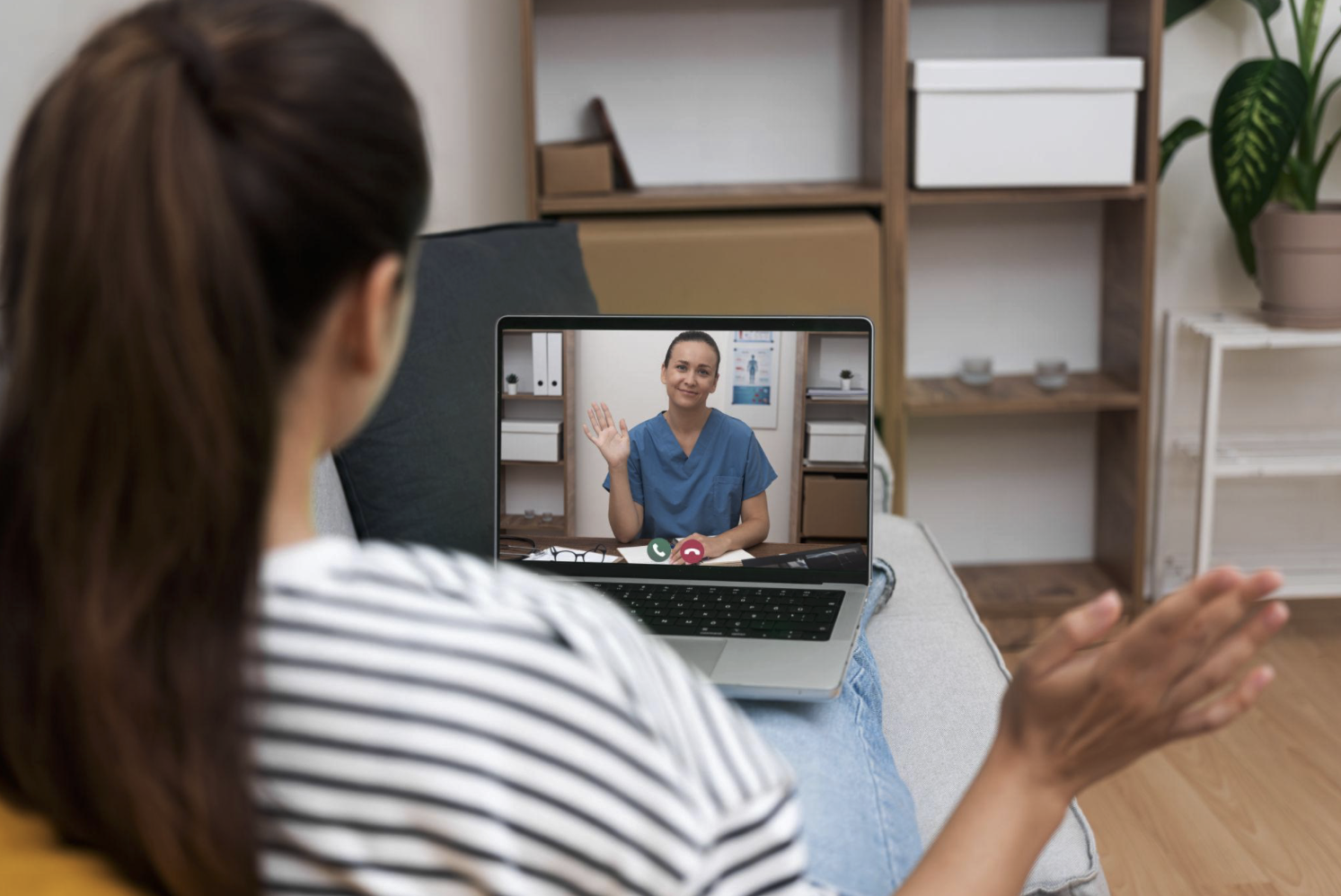 A woman on a video call with a nurse, waving hello, on her laptop while sitting on a couch.