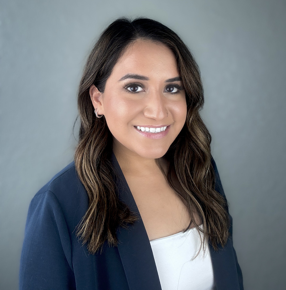 Portrait of a smiling woman with long, wavy brown hair, wearing a navy blazer over a white top, and earrings, against a gray background.