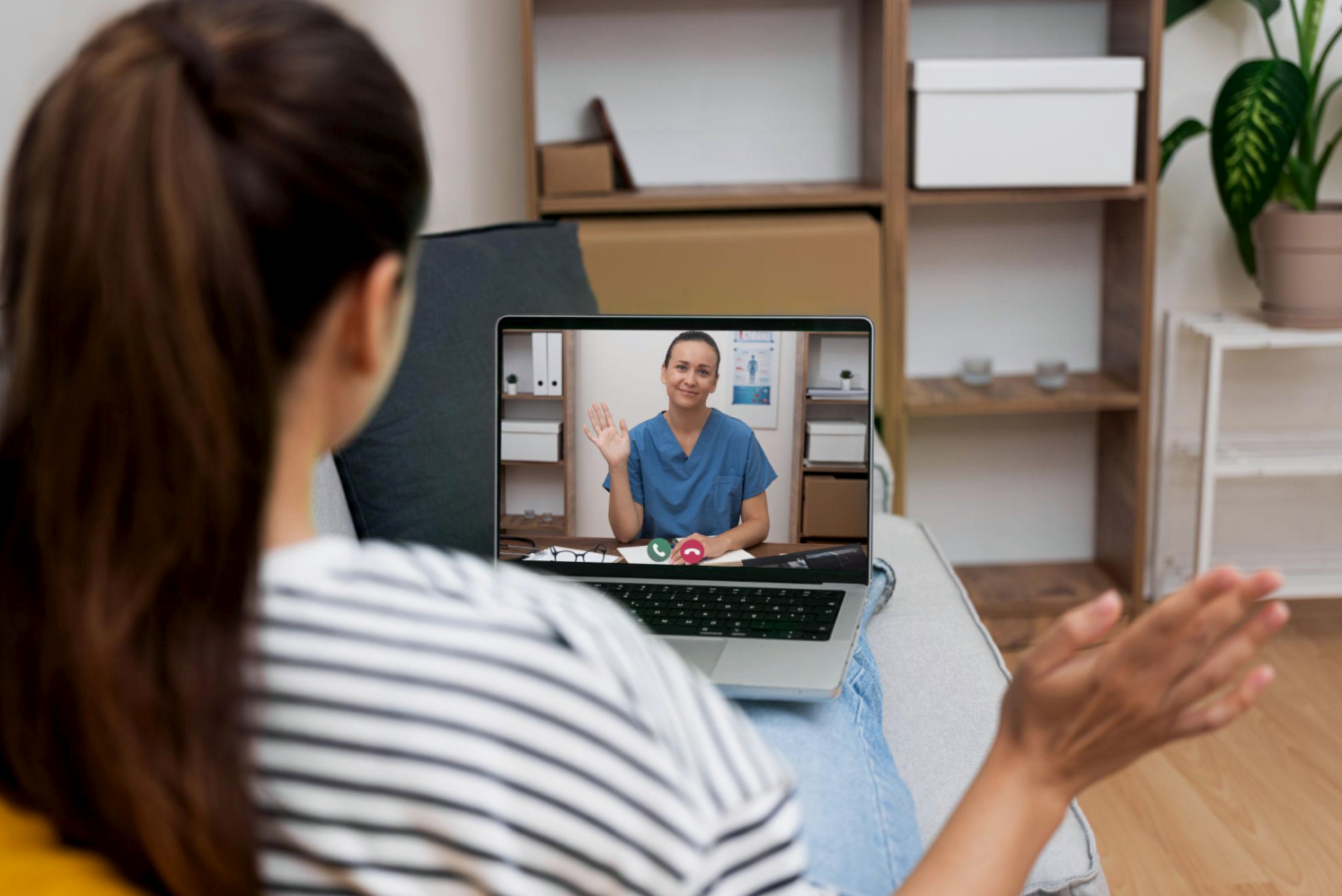 A woman with brown hair wearing a striped shirt is sitting on a couch, engaging in a video call with a female healthcare professional on her laptop screen, waving hello.