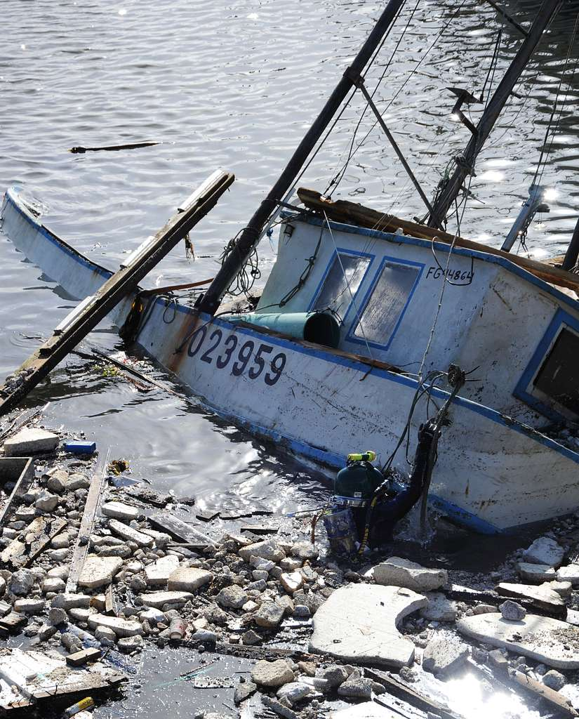 Sailboat partially submerged and wrecked on a rocky shoreline, with debris and broken parts around it.