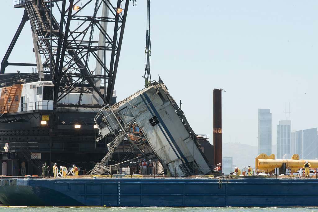 A large oil rig with a crane is in the process of dismantling with the destroyed crane hanging down. Workers are present on the site, and a city skyline with tall buildings is visible in the background.