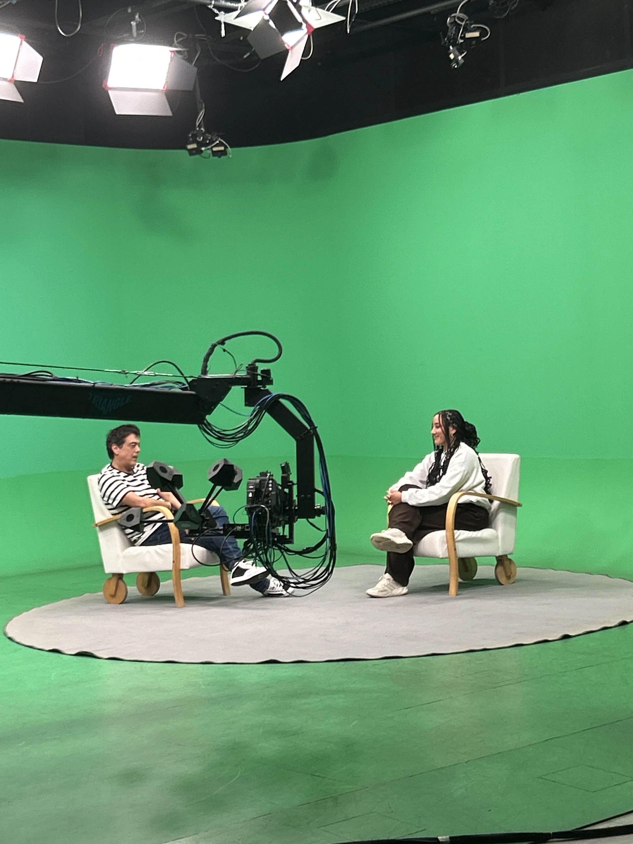 Two people sitting on chairs in front of a green screen, with professional studio lighting and camera equipment.