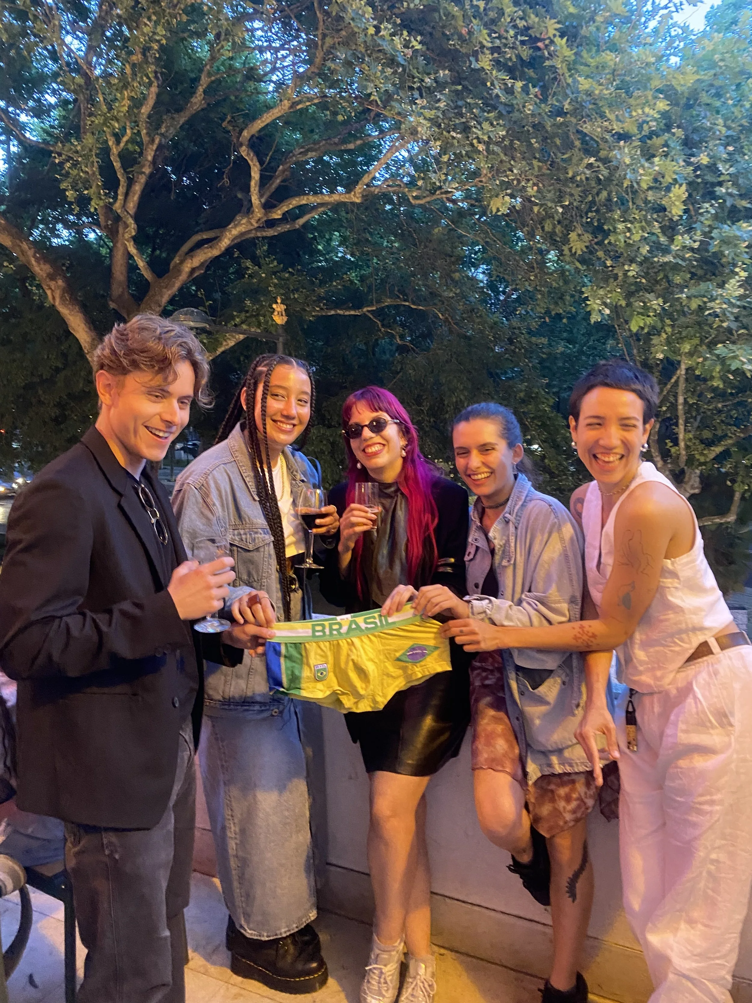 A group of six friends at an outdoor gathering, smiling and holding drinks, standing under a large tree during the evening.