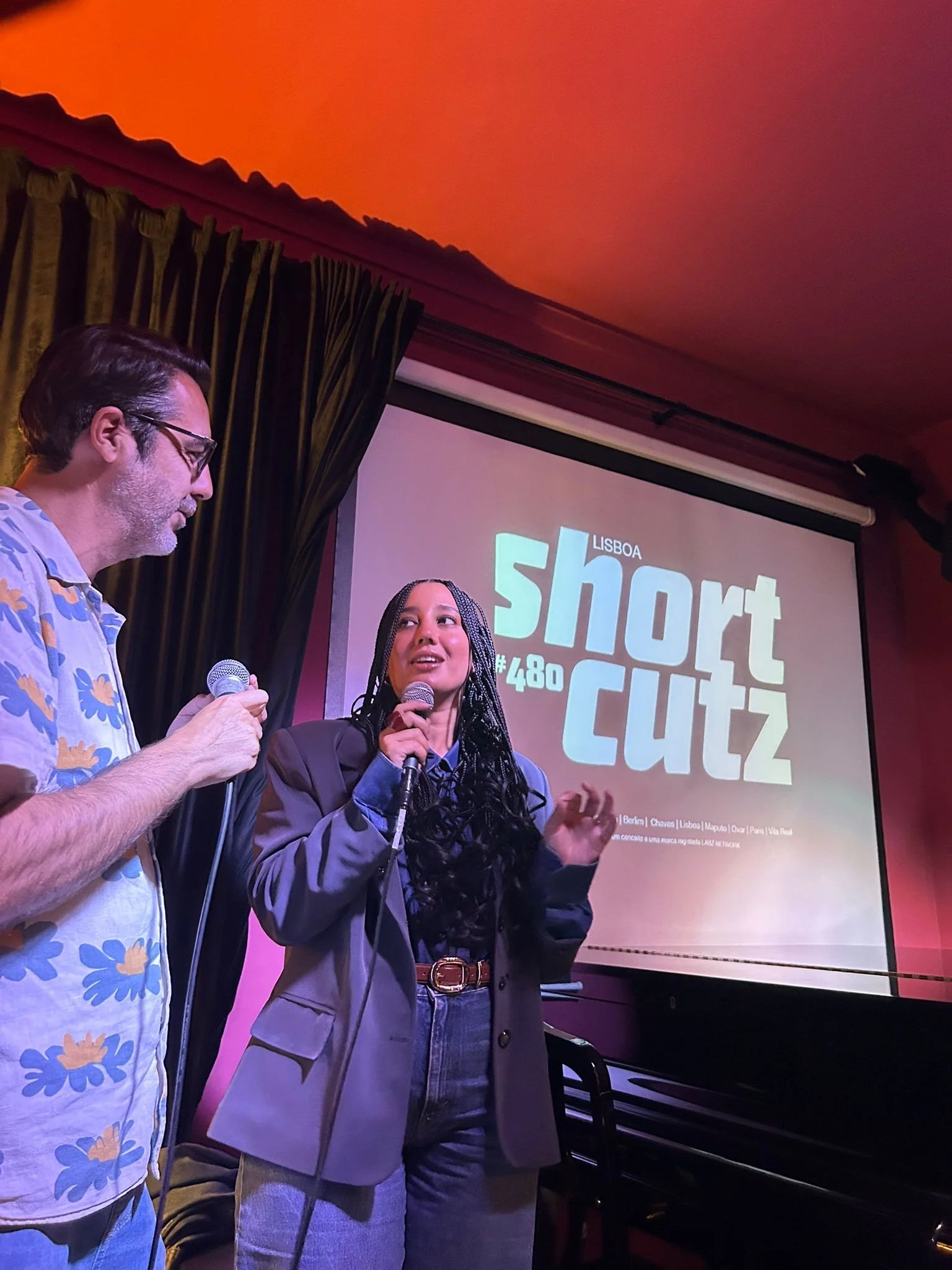 Two people speaking at an event, with a women holding a microphone and a man with glasses holding a microphone, in front of a presentation screen displaying event details in Lisbon.