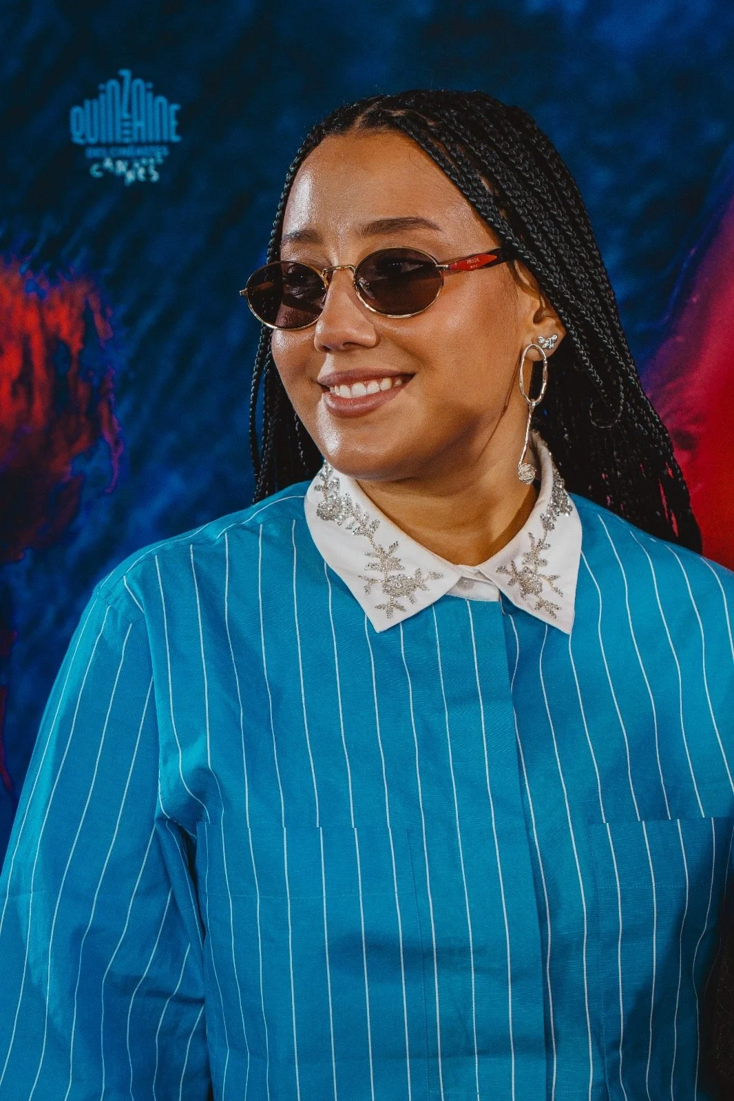 A woman with braided hair wearing sunglasses, hoop earrings, a white collared shirt with silver snowflake embroidery, and a blue pinstripe blazer, smiling against a colorful abstract background.